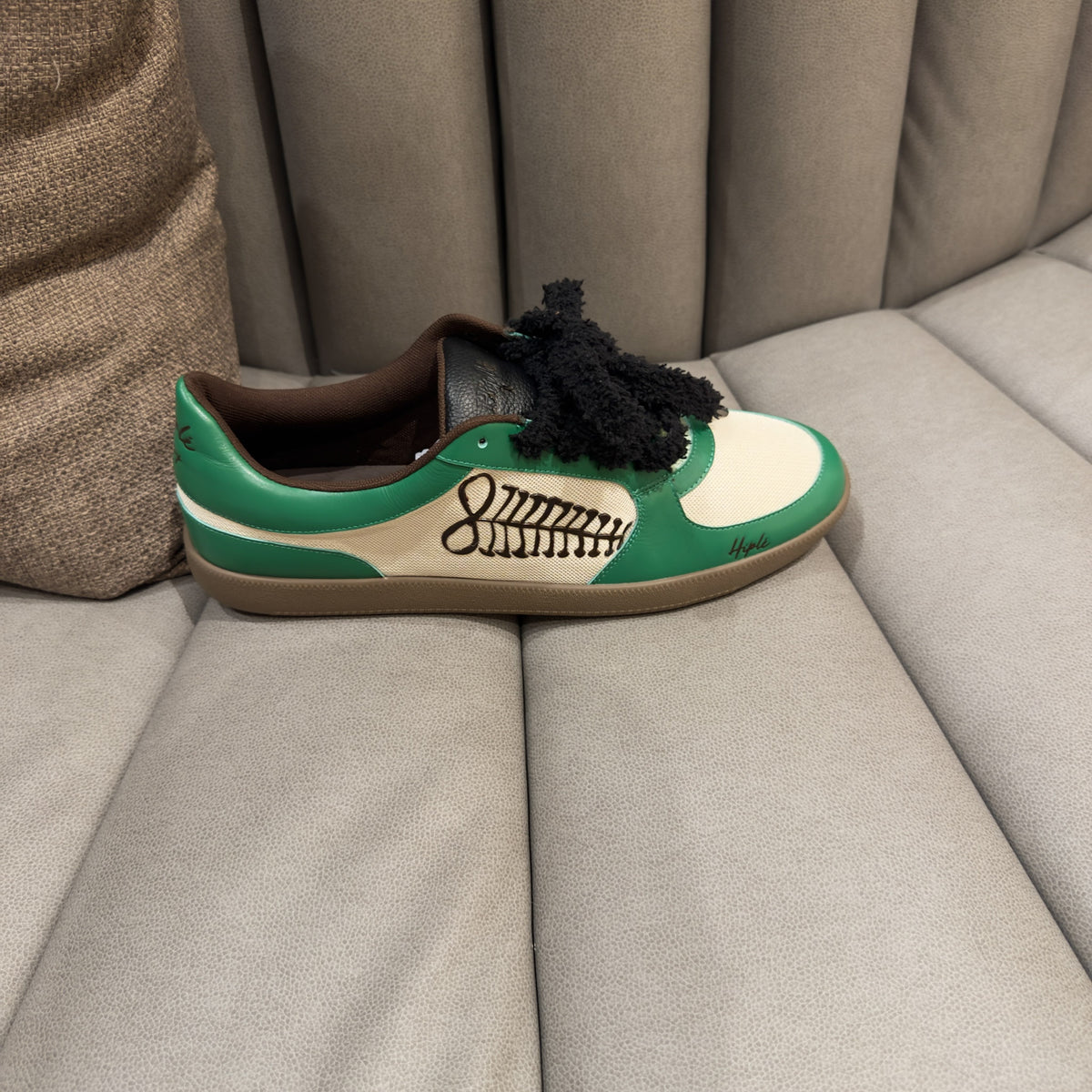 A green and off-white low-top sneaker resting on a light gray upholstered sofa. The sneakers are the ayas sneakers by hiplé