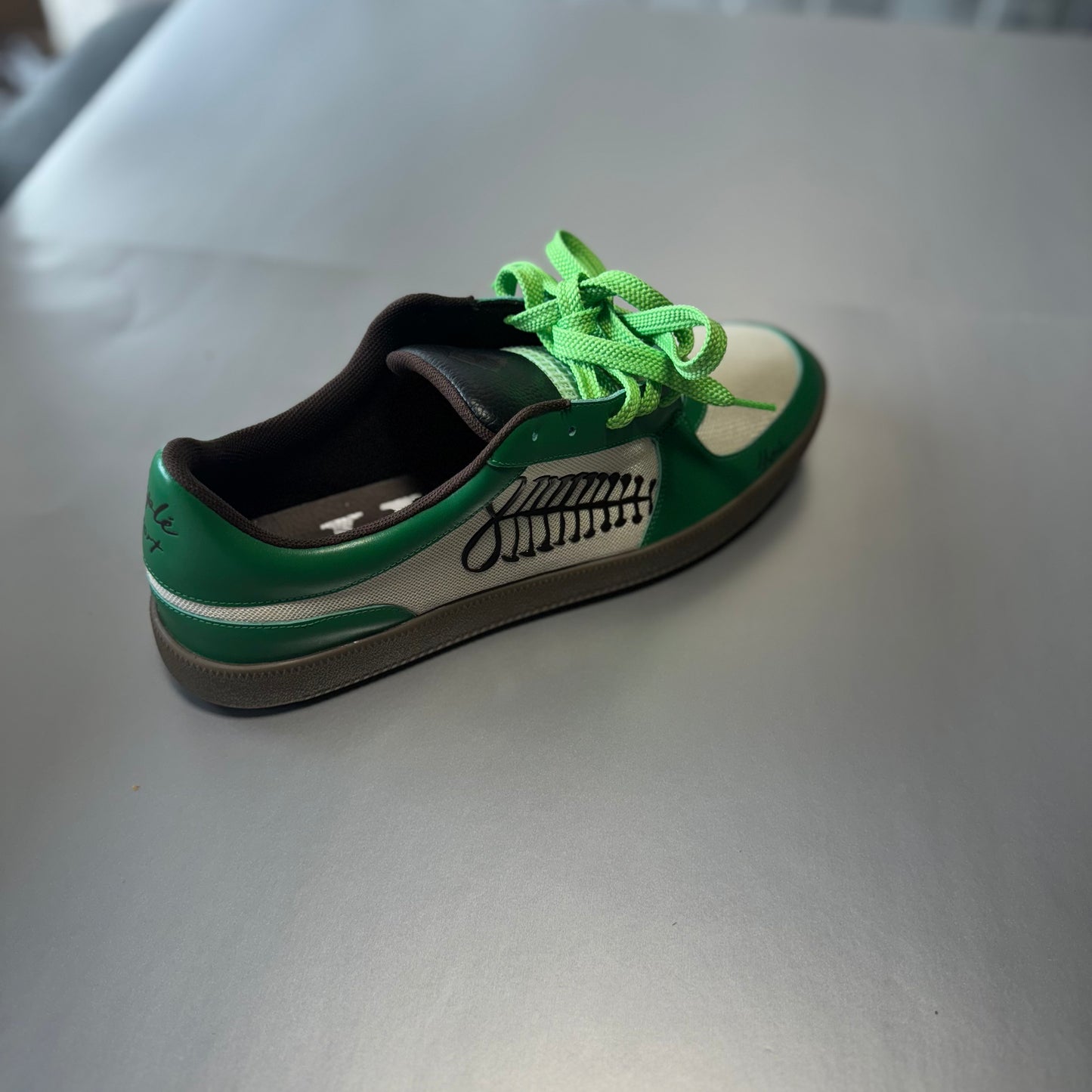 angled view of a green and white low-top ayas sneaker by Hiplé with bright neon green laces, a black graphic logo on the side, and a brown gum sole, resting on a gray surface. (hiple)