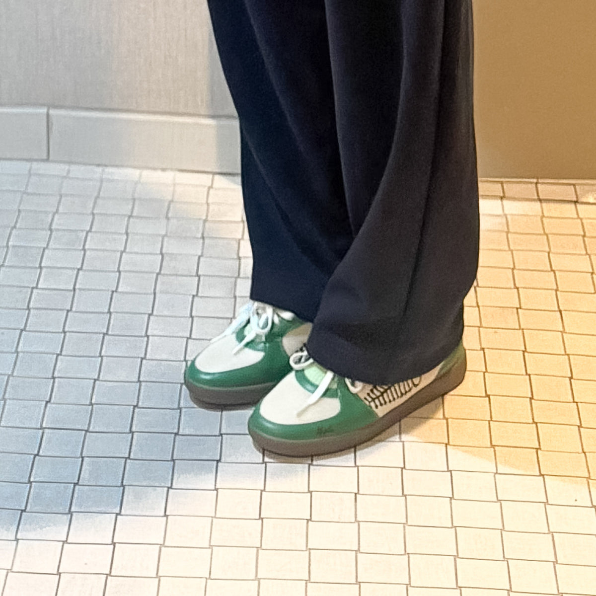 A person wearing dark navy wide-leg pants and sb green and white low-top sneakers known as the ayas while standing on a white tiled floor. (Hiplé)