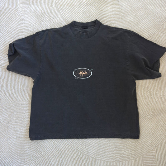 A grey garment dye short-sleeve t-shirt with a small white and gold 'Hiplé' logo printed on the chest, laid flat on a textured beige fabric background. By the brand Hiplé (Hiple)