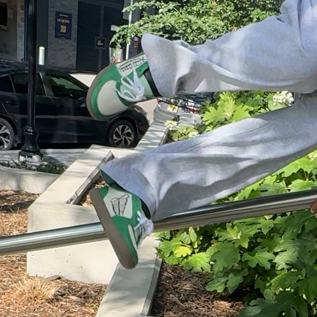 A action product photo showcasing the ayas sneaker by hiple. A person outdoors wearing light gray sweatpants and green-and-white sneakers while balancing on a metal railing near green plants (Hiplé)